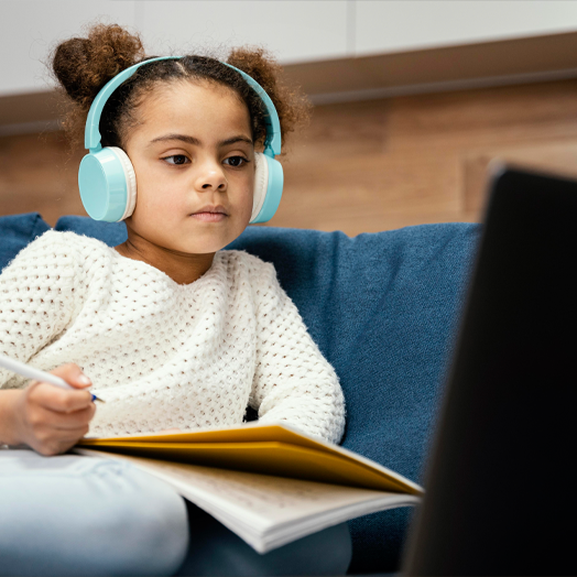 Young girl in K-5 learning synchronously with blue headphones on and a notebook in hand, holding a pen for notes